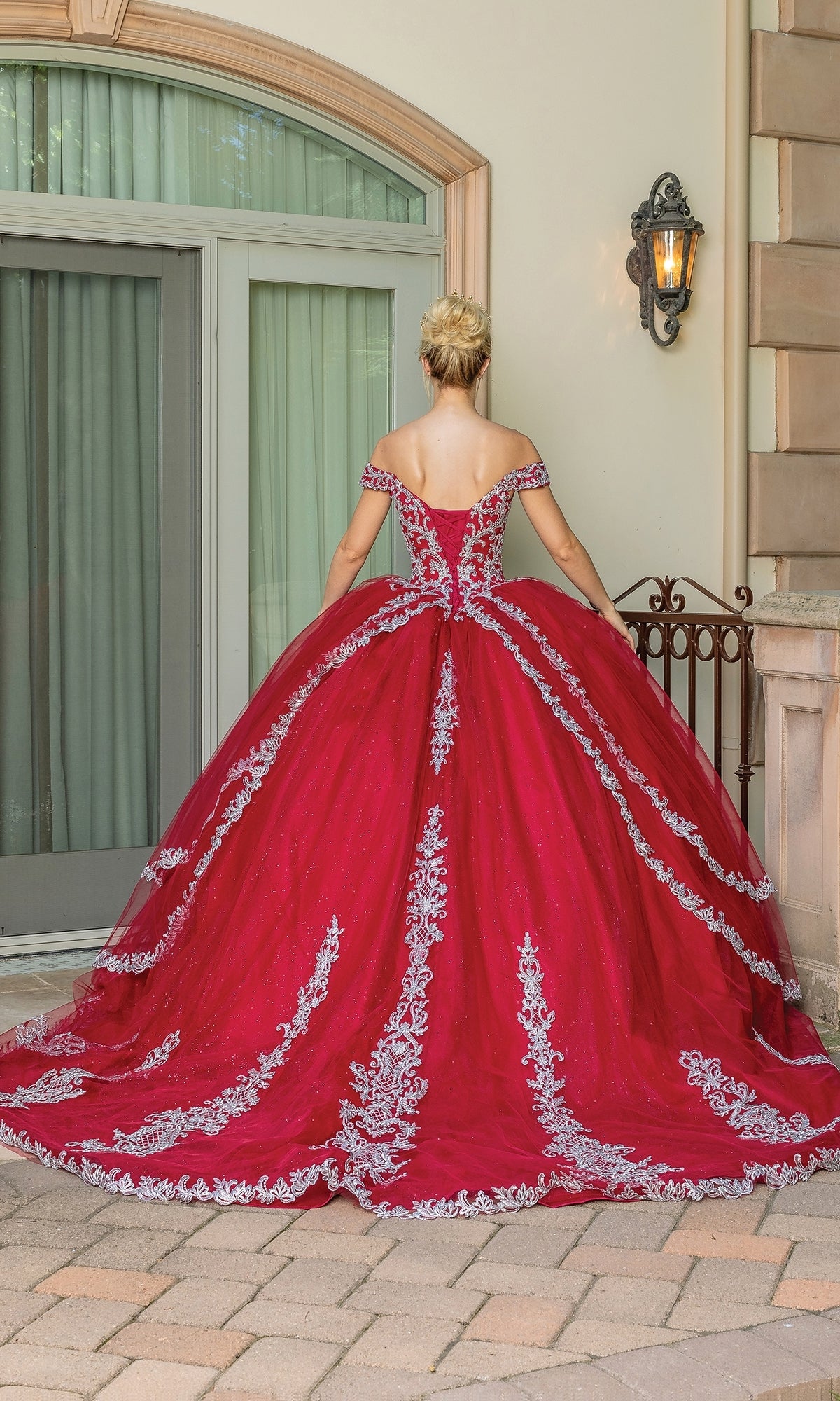 A woman faces away, wearing the Red Off-the-Shoulder Quinceanera Dress 1662—burgundy with a sweetheart neckline and silver embroidery—outside near a building with large windows and a lantern.